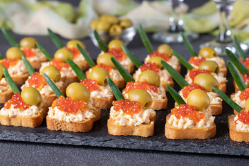 Festive canapes with red caviar and olives on croutons of white bread on slate board on gray background, Closeup
