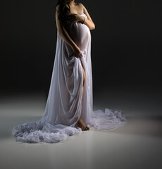 Fototapeta na wymiar A pregnant woman is standing in a white dress on a black background. Studio pregnancy photo shoot. Space for text. Woman in white dress.