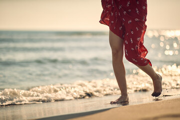 Shot of legs of young woman in red summer dress walking on beach by waves of water. Fashion,...