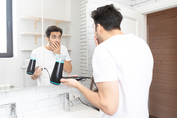 Funny man standing in front of the mirror and amusing with hair dryer in the bathroom during daily morning routine. Bad hair day concept