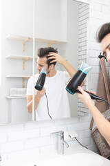 Funny man standing in front of the mirror and amusing with hair dryer in the bathroom during daily morning routine. Bad hair day concept