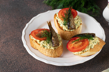 Homemade sandwiches with sprats, egg, cheese and tomatoes on white plate on brown table