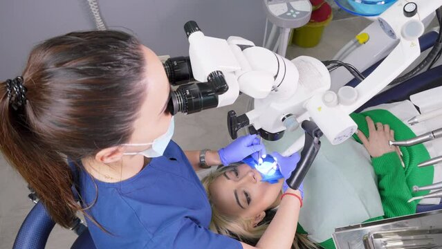 Female dentist using dental microscope treating patient teeth dental clinic office. undergoing treatment by experienced dentist using microscope. Image of woman on patient's chair in dentist's office.