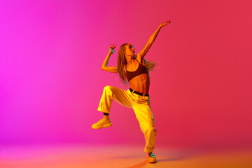 Naklejka na ściany i meble Portrait of young stylish woman, hip-hop dancer training in casual clothes over gradient pink background in neon light. Street style dance