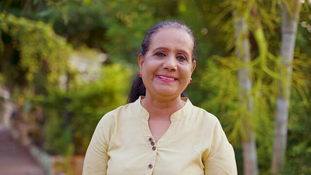 Happy smiling indian senior woman standing by looking camera at park - concept of retirement lifestyle , successful and relaxation