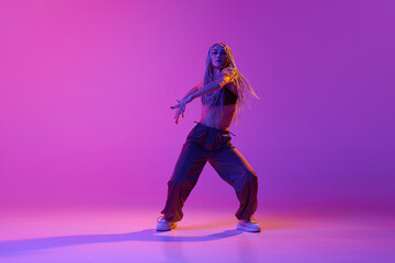 One professional hip-hop dancer wearing fashion clothes moving with inspiration over gradient purple background in neon light. Modern choreography