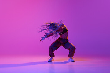 Fototapeta na wymiar Portrait with one young girl, inspired dancer with pigtails dancing with hands over gradient purple background in neon light. Flying braided dreads