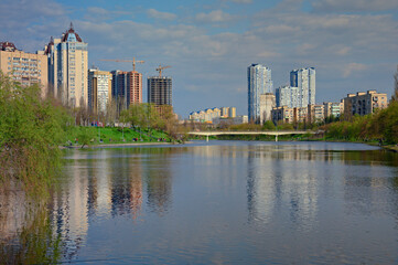 Wide-angle landscape view of water channel near Rusanivka neighborhood. High water in Kyiv, spring 2023, Ukraine. Residential high-rise buildings in the background against blue sky. Flooding