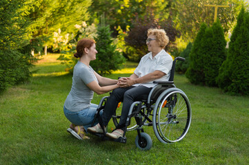 A caucasian woman holds the hand of an elderly mother sitting in a wheelchair. Walk outdoors. 