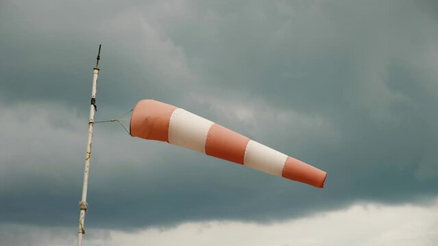 A white and red windsock on a windy day, mounted on a pipe with peeling paint. Against a dark sky with a huge cloud. Close-up