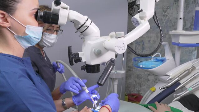Two women dentist working with dental microscope. Doctor and his assistant in dental clinic. Doctor looking through microscope and holding dental probe and mirror. Modern dental clinic. Dental office.