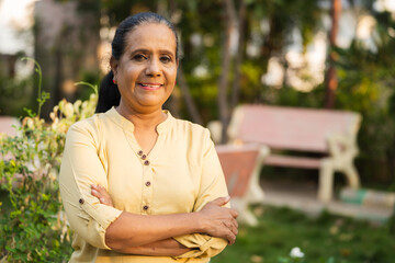 Happy smiling indian senior woman confidently standing with arms crossed by looking camera at park - concept of retirement lifestyle , successful and relaxation