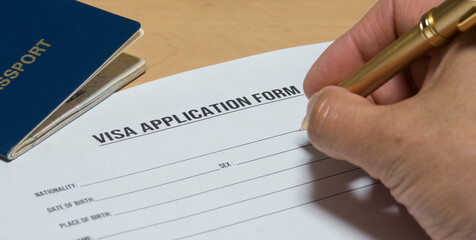 hand holding a pen writing a visa application form. Form and passport on the desk.