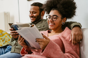 Cheerful woman reading book while sitting on couch with indian man who's using mobile phone