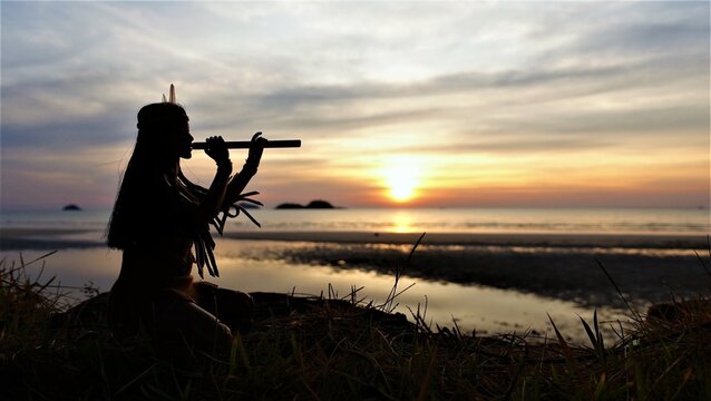 A Native American Indian plays a bamboo flute. Silhouette against the background of sunset.