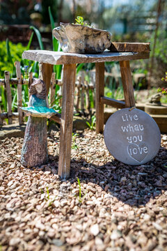 A small wooden figure holds a blue leaf and stands next to a wooden stool. there is a sign next to it that says: live what you love, in the garden