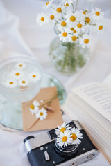 Chamomile flowers in vase, vintage camera and book on white fabric.