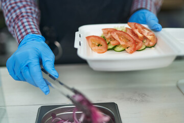 The cook preparing healthy vegetarian lunch box for take away. Kitchen worker take vegetables with tongs and put them in a white styrofoam container