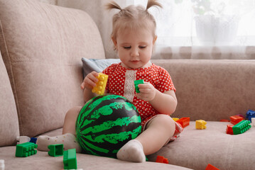 Portrait of cute baby in polka dot dress sitting on sofa with ball and colorful blocks. Kid playing toys in living room.