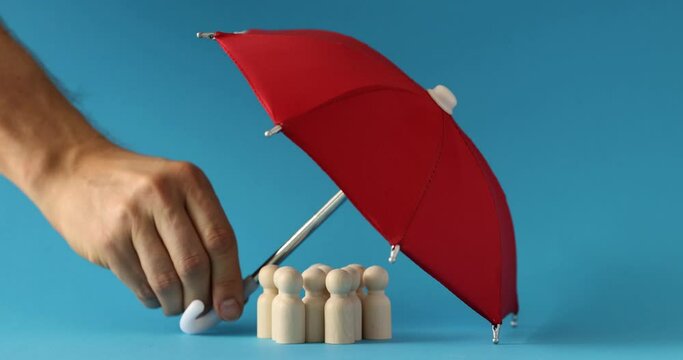 Male hand of insurance agent putting red umbrella over wooden men on blue background 4k movie slow motion. Social assistance to poor citizens concept