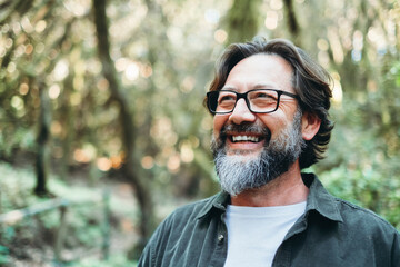 Portrait of young mature man alone smiling outside with nature trees in background. Environment. Happy people enjoying outdoor leisure activity. Nature feeling. Bearded adult male wearing eyeglasses - obrazy, fototapety, plakaty