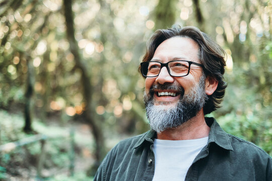 Portrait of young mature man alone smiling outside with nature trees in background. Environment. Happy people enjoying outdoor leisure activity. Nature feeling. Bearded adult male wearing eyeglasses