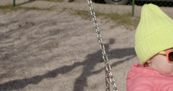 The balance of the child's psyche, calmness. She moves forward and backward on a sunny day. Relaxation of the child in free time. Toddler girl in glasses and a hat rides on a swing in the yard. 