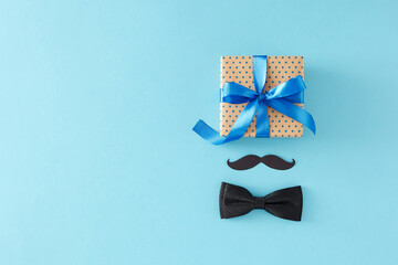 Stylish Father's Day gift concept. Top view flat lay of gift box black bow tie and paper mustache on light blue background with empty space for greeting text or advert