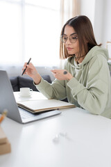 Brunette freelancer in eyeglasses holding smartphone near laptop and notebook on table at home.
