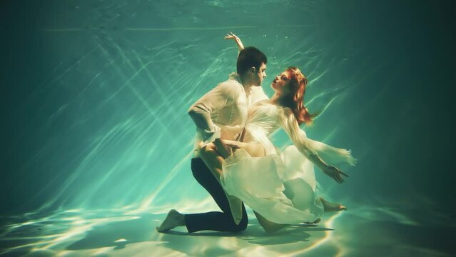 Fantasy couple fall in love man king and woman fashion model posing underwater dark blue sea. Wet white dress. Fairy tale sexy girl Greek goddess Muse river swims dancing spinning guy under water pool