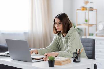 Brunette freelancer using laptop near notebooks and books at home.