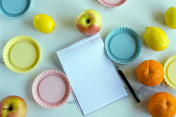 An open paper notebook and pen on a white kitchen table among fresh fruits and a variety of nuts. The concept is the compilation of a menu, a list of products, and the design of recipes. copy space