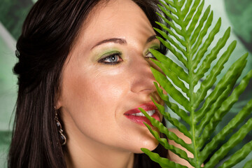 Naklejka premium Half covered face close-up. Beautiful brunette woman with fresh green makeup enjoying the floral environment, posing among plants with big tropical leaves. Respecting the nature concept.