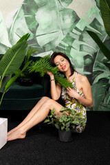 Obraz premium Beautiful brunette woman with fresh green makeup enjoying the floral environment, posing among plants with big tropical leaves. Respecting the nature concept.