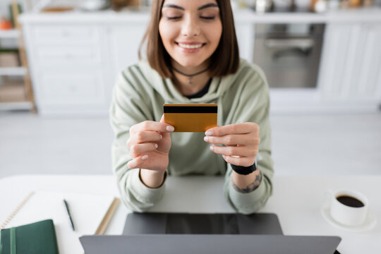 Smiling woman holding credit card near laptop and coffee on table at home.