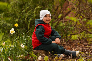 A little boy in spring clothes sits on a tree stump among flowers and smiles.