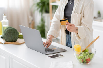 Fototapeta premium Cropped view of tattooed woman holding credit card and using laptop near blurred salad in kitchen.