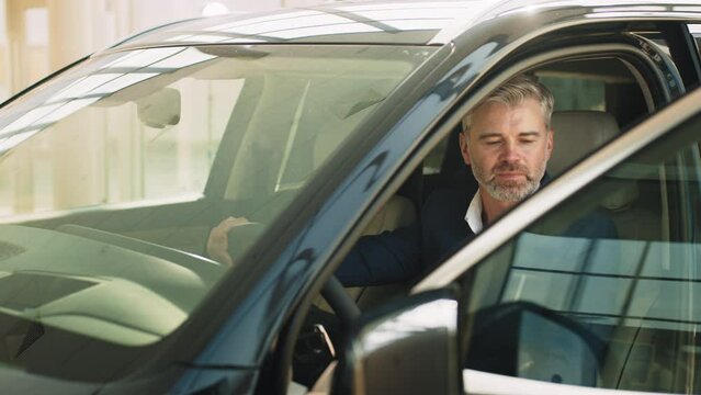 Portrait of a handsome beard grey hair mature man sitting in a brand new car at the dealership. An adult man driving a motor vehicle on a sunny day, captured in a headshot portrait through the window.