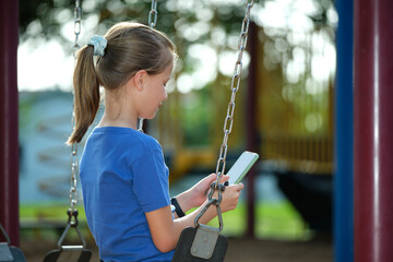 Teen child girl using cellphone sitting on swing in park during summer vacations