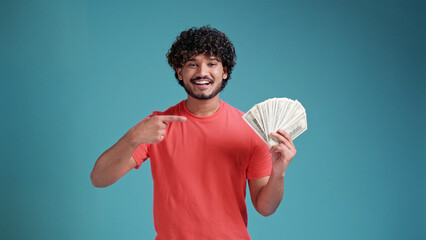 Smiling cheerful happy young bearded latin spanish man 20s years old showing fan of cash money in dollar banknotes looking camera isolated on plain blue background studio portrait