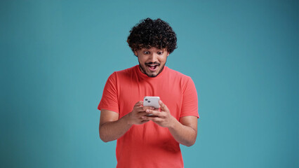 young latin man looks delighted into smartphone and says wow. Dressed in casual on a blue studio...