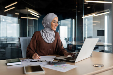 Muslim young woman in hijab programmer, developer working in office behind notebook and with documents. - obrazy, fototapety, plakaty