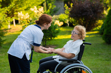 A nurse holds an elderly caucasian woman in a wheelchair by the hand as support. Nurse walks with a patient in the park. 