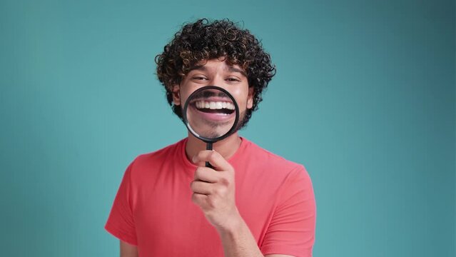 funny latin spanish man shows white teeth and smiles through a magnifying glass on blue studio background