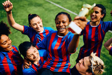 African American soccer player holds winning trophy while being carried by her teammates at stadium.