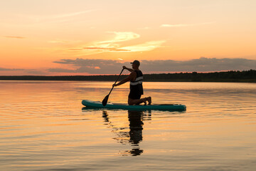 A man on a SUP board on his knees with an oar at sunset against a pink sky floats in the water of the lake.