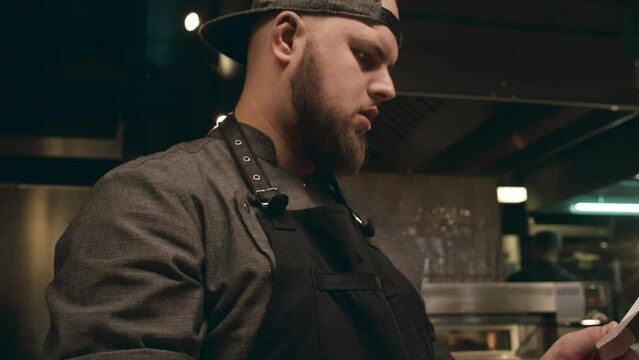 Medium close-up follow shot of young bearded Caucasian male chef in apron and cap taking order sheet in restaurant kitchen, reading it, then beginning to prepare fresh vegetables