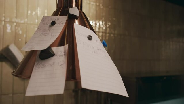 Close-up lockdown shot of metal lamp in restaurant kitchen, paper slips with orders hanging on magnets, and tattooed hands of male chef taking off one sheet