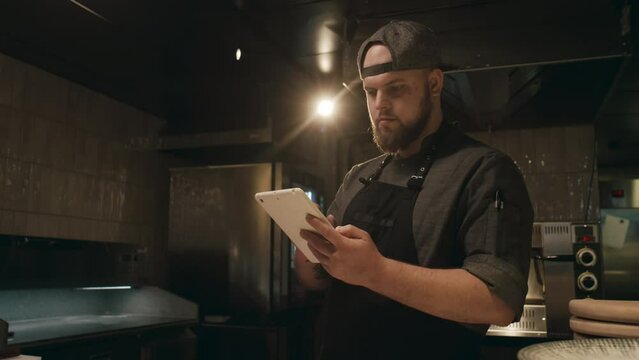 Medium shot of young bearded Caucasian man in chef uniform standing in dimly lit restaurant kitchen, waiting for orders and using tablet computer