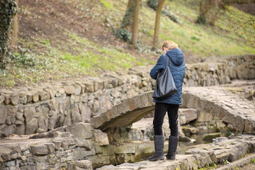 A girl stands thoughtfully near a stone bridge on the bank of a stream in a park with green spaces....